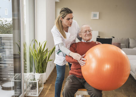 Hauspflegerin unterstützt älteren Mann bei Übungen mit dem Fitnessball - UUF31262