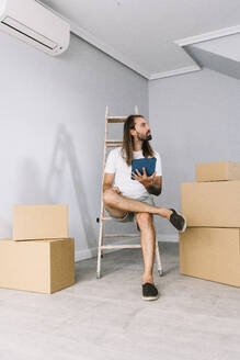 Junger Mann sitzt auf einer Leiter mit einem Tablet-PC in der Nähe von Kartons in seinem neuen Zuhause - DAMF01310