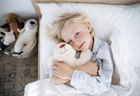 Lächelnder Junge liegt mit Teddybär auf dem Bett zu Hause - NJAF00765
