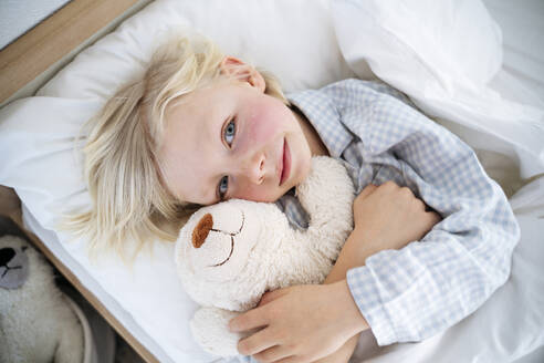 Lächelnder Junge umarmt Teddybär auf Bett zu Hause - NJAF00766
