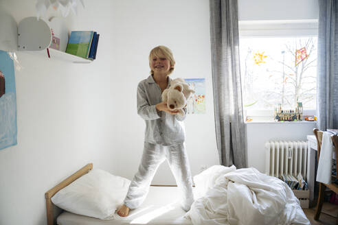 Glücklicher Junge hält Teddybär und steht auf dem Bett zu Hause - NJAF00769