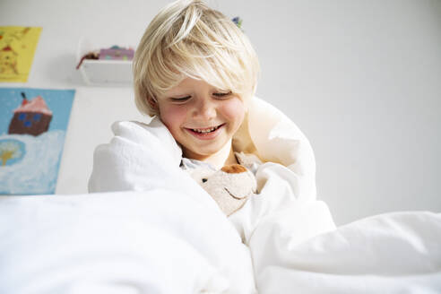 Happy boy playing with teddy bear wrapped in blanket at home - NJAF00771