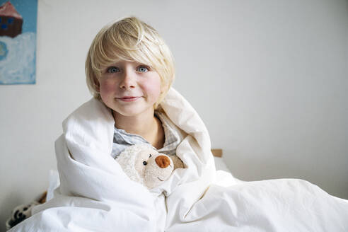 Blond boy with teddy bear wrapped in blanket at home - NJAF00772