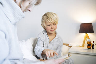 Junge, der auf ein Buch zeigt, das mit seiner Mutter im Schlafzimmer liegt - NJAF00784