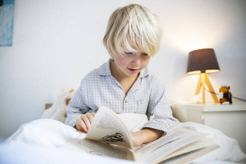 Blond boy reading book sitting on bed at home - NJAF00785