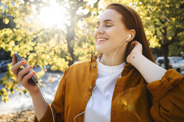 Smiling woman listening to music through wired in-ear headphones in autumn - ALKF00963
