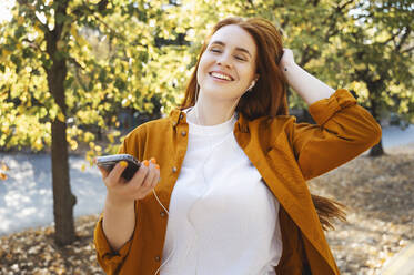 Happy woman with hand in hair listening to music in autumn - ALKF00965