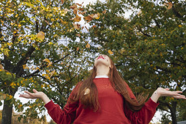 Rothaarige Frau spielt mit Herbstblättern unter einem Baum im Park - ALKF00972