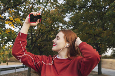 Carefree woman enjoying listening to music through wired in-ear headphones at park - ALKF00979