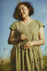 Mature woman holding dandelion and standing in field on sunny day - IEF00539