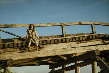 Unbekümmerte Frau sitzt auf einer Holzbrücke unter blauem Himmel - IEF00540