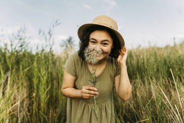 Playful woman wearing hat and holding dandelion near face in field - IEF00562