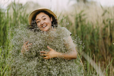 Fröhliche reife Frau, die einen Strauß Schleierkrautblüten auf einem Feld umarmt - IEF00583