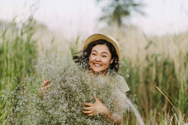 Cheerful woman wearing hat and holding bunch of gypsophila flowers in field - IEF00588