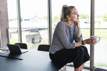 Businesswoman holding coffee cup and sitting on desk in office - UUF31275