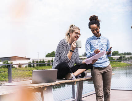 Smiling businesswoman sitting on bench and having discussion with colleague - UUF31284