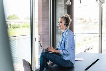 Smiling businesswoman sitting on desk with laptop in office - UUF31290