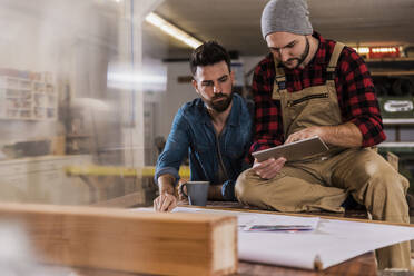 Tischler teilt sich einen Tablet-PC mit einem Kollegen, der auf einer Werkbank in der Werkstatt sitzt - UUF31297