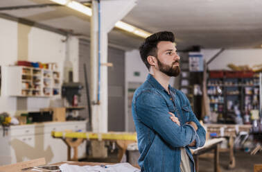 Thoughtful carpenter standing with arms crossed at workshop - UUF31302