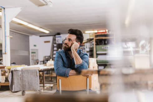 Young carpenter sitting on chair and day dreaming at workshop - UUF31306