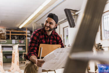 Smiling carpenter holding blueprint sitting on chair at workshop - UUF31308