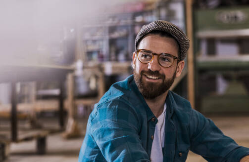Lächelnder Schreiner mit Brille und flacher Mütze in der Werkstatt - UUF31310