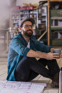 Lächelnder Tischler mit Blaupause und Kaffeetasse in der Hand in der Werkstatt - UUF31311