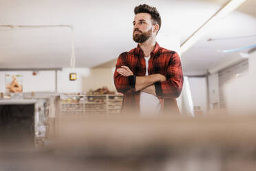 Thoughtful carpenter with arms crossed standing at workshop - UUF31321