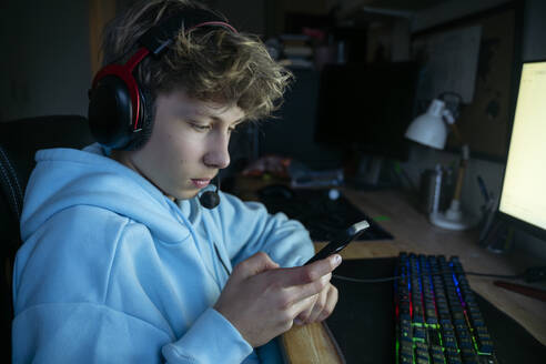 Junge sitzt in der Nähe eines Computers und benutzt ein Smartphone zu Hause - NJAF00793