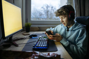 Jugendlicher, der zu Hause am Computer sitzt und sein Smartphone benutzt - NJAF00794