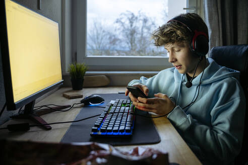 Teenage boy sitting near computer and using smart phone at home - NJAF00794