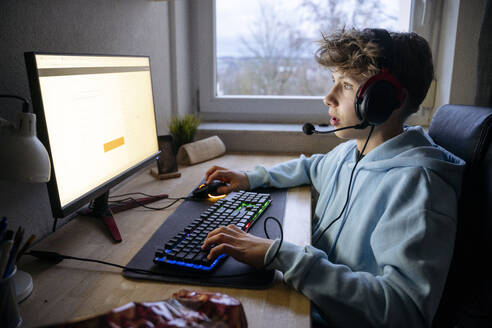 Junge trägt ein Headset und spielt ein Videospiel auf einem Computer am Schreibtisch - NJAF00797