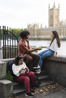 Woman using smart phone with friends sitting near Thames river in London city - ASGF04880