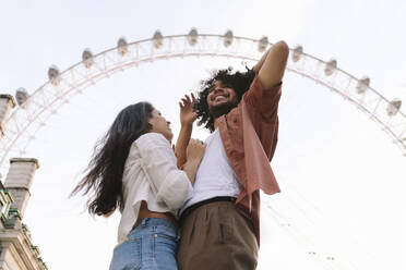 Happy couple standing in front of Ferris wheel under sky in London - ASGF04885