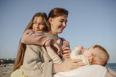 Happy woman playing with baby girl near daughter at beach on sunny day  - ALKF01002