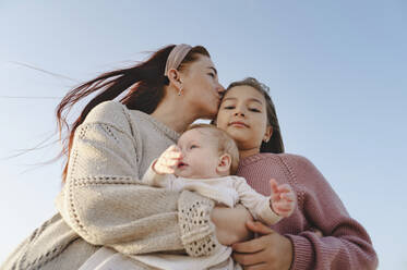 Woman holding baby girl and kissing daughter under clear sky - ALKF01006