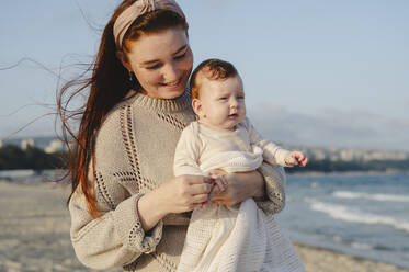 Smiling mother holding daughter at beach on sunny day - ALKF01009