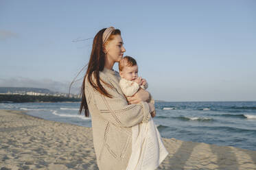 Thoughtful woman holding daughter and standing near sea at beach - ALKF01011