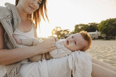 Smiling woman sitting with daughter on lap at beach - ALKF01030