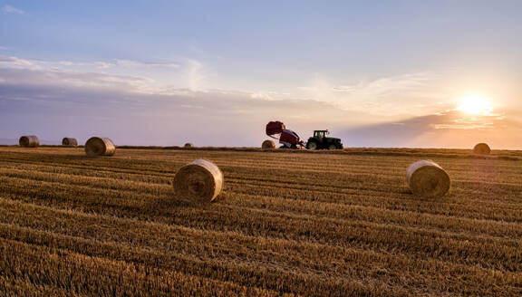 Tractor baling agricultural field under sky at sunset - NOF00928