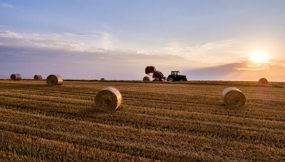 Traktor beim Pressen von Ballen auf einem landwirtschaftlichen Feld unter dem Himmel bei Sonnenuntergang - NOF00928