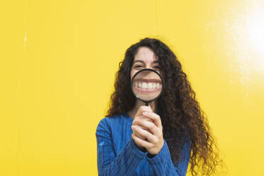 Frau hält Vergrößerungsglas in der Nähe des Mundes vor gelbem Hintergrund - MGRF01064