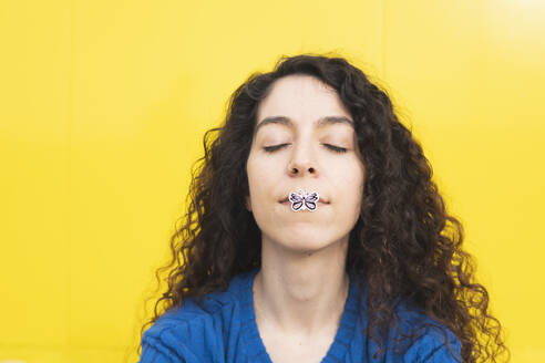 Frau mit Schmetterlingsaufkleber auf der Lippe vor gelbem Hintergrund - MGRF01079