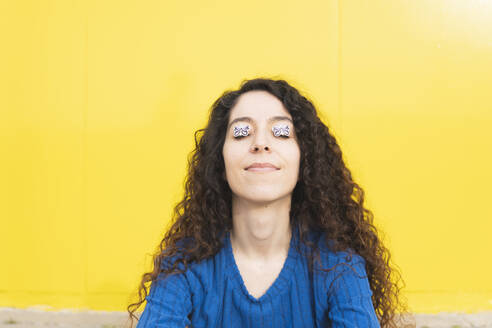 Frau posiert mit Schmetterlingsaufklebern im Gesicht in der Nähe einer gelben Wand - MGRF01083