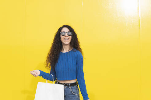 Glückliche Frau mit Sonnenbrille und Einkaufstüten vor gelbem Hintergrund - MGRF01088