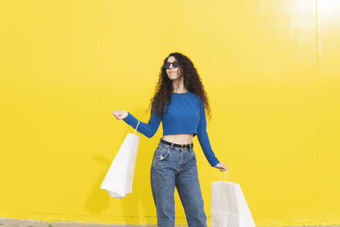 Frau mit Einkaufstüten vor einer gelben Wand stehend - MGRF01089