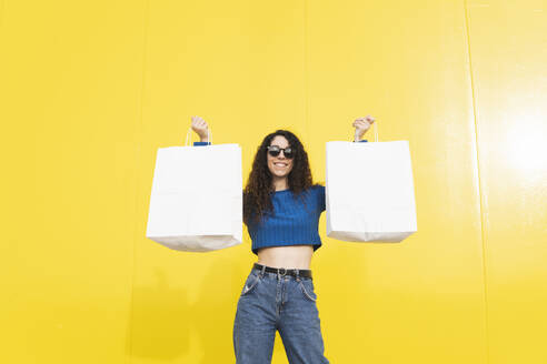 Glückliche Frau posiert mit Einkaufstüten vor gelbem Hintergrund - MGRF01091