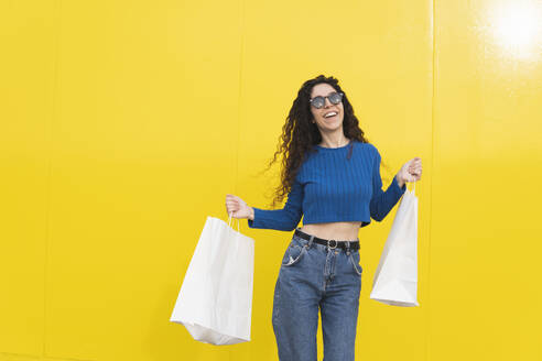 Glückliche Frau tanzt mit Einkaufstüten vor gelbem Hintergrund - MGRF01093