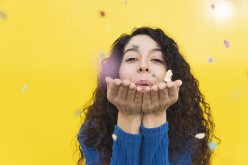 Woman blowing colorful confetti against yellow background - MGRF01094