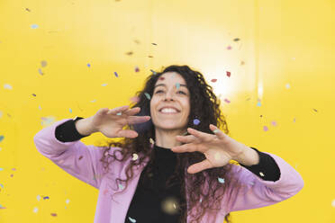 Smiling woman playing with confetti against yellow background - MGRF01108